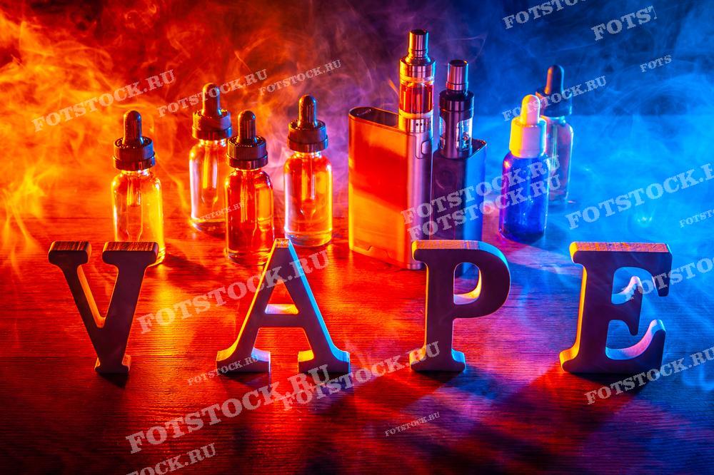 На фотографии изображены буквы, составляющие слово "VAPE", и различные аксессуары для вейпинга, такие как флаконы с жидкостью и электронные устройства, расположенные на деревянной поверхности. Фон украшен разноцветным дымом, создающим эффектный визуальный контраст.